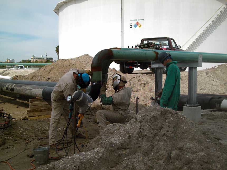 ACS crew welding a green-coated pipeline beside a white fuel storage tank