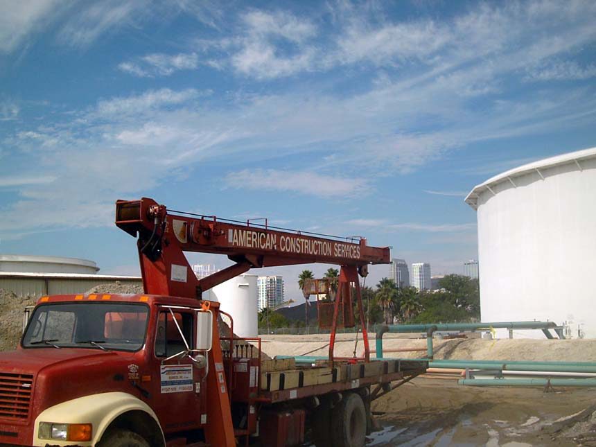 Red American Construction Services crane truck at a Tampa fuel terminal with the downtown skyline behind